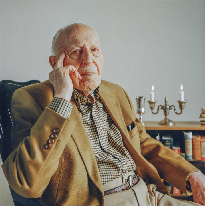 A man sits in a chair with his hand to his face, next to a lit candelabra and books.
