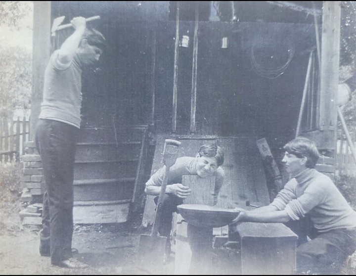 A man swings an axe while two other men work with a bowl and wood in front of a wooden building.