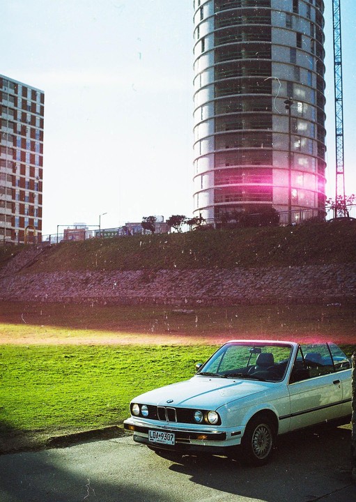 A white convertible car is parked on a paved surface next to a grassy hill with modern buildings in the background.
