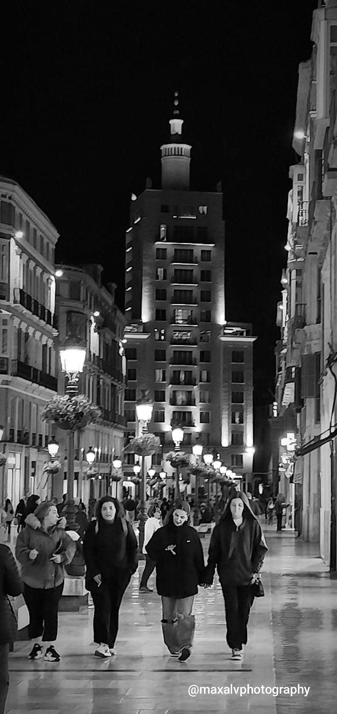 Three women walk down a brightly lit street at night with tall buildings on either side.