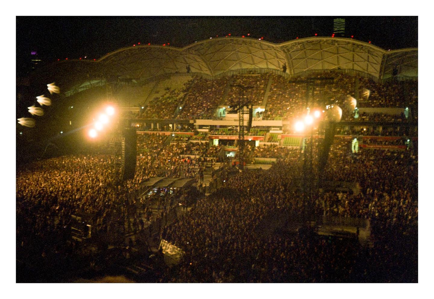 A large crowd of people are gathered in a stadium at night, illuminated by bright stage lights.