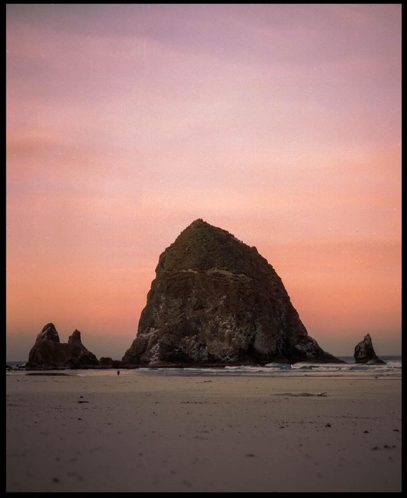 A large rock formation sits on a sandy beach under a colorful sky at dusk.
