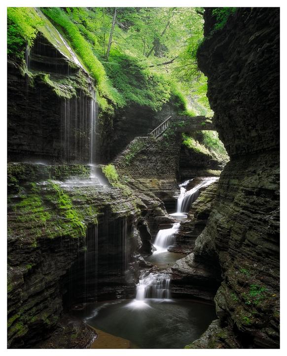 A series of waterfalls cascade down a mossy gorge with a stone bridge and stairs visible in the background.