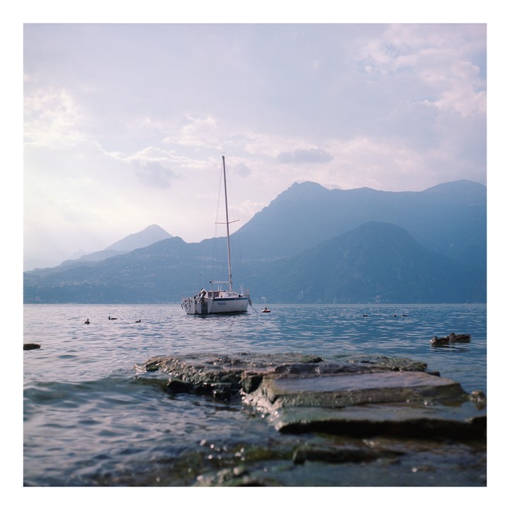 A sailboat floats on a lake with mountains in the background and rocks in the foreground.