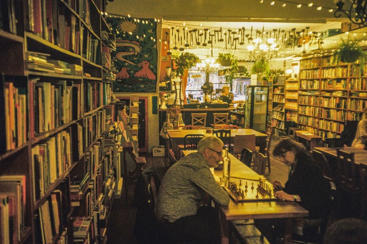 A man and a woman play chess at a table in a bookstore with many shelves of books and string lights.