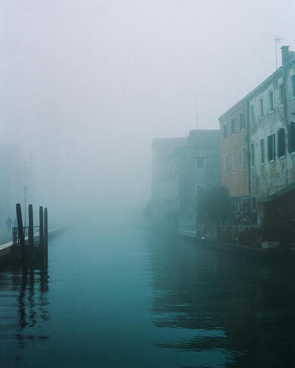 A canal in Venice is shrouded in thick fog, with buildings visible on the right and mooring posts on the left.
