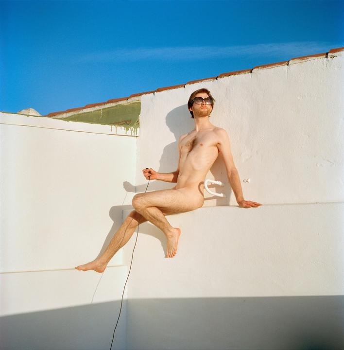 A man wearing sunglasses sits nude on a white wall under a clear blue sky.