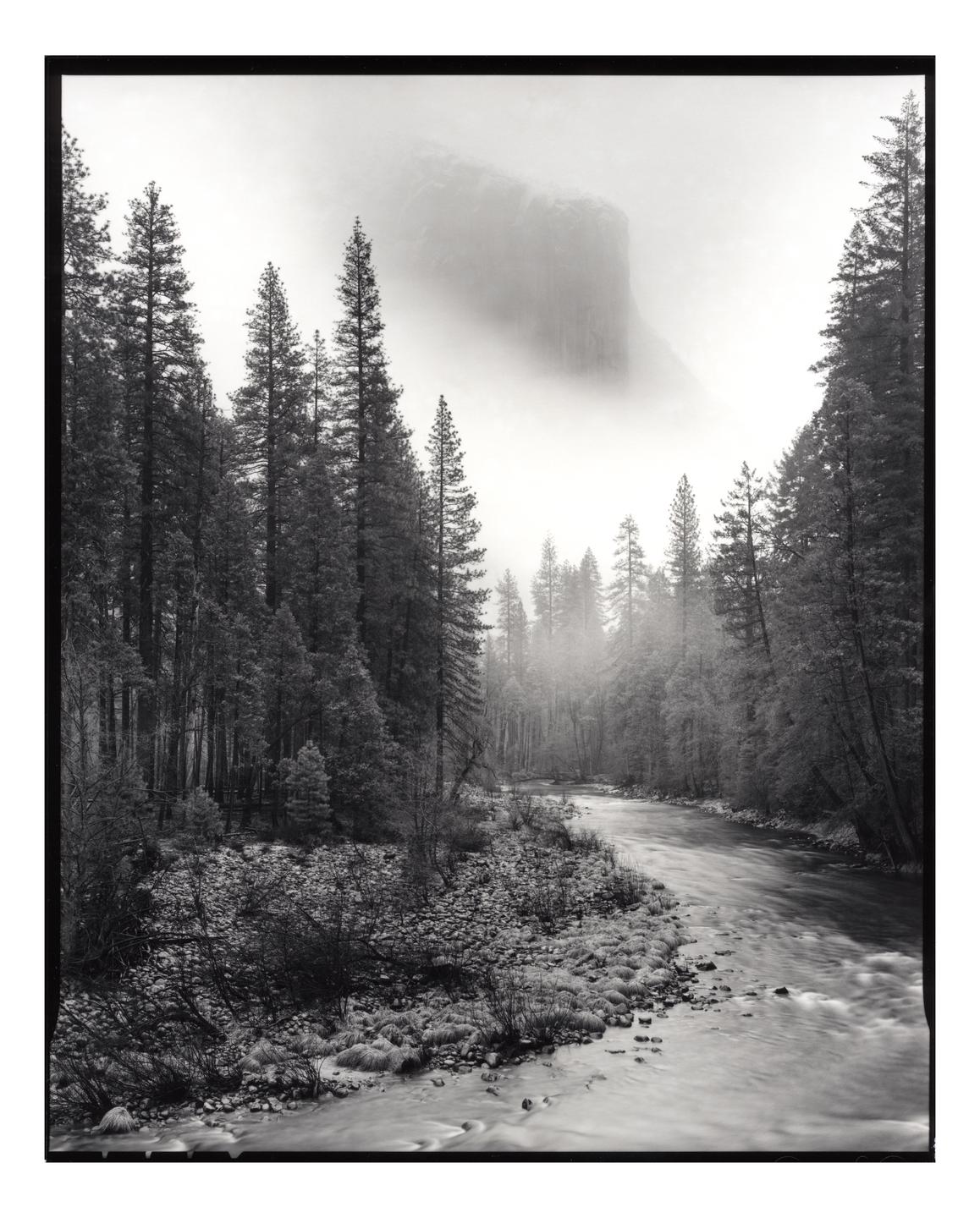 A river flows through a forest with a large rock formation in the background shrouded in fog.