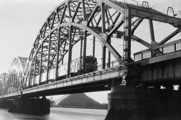 A train crosses a large metal bridge over water, with a building visible in the background.