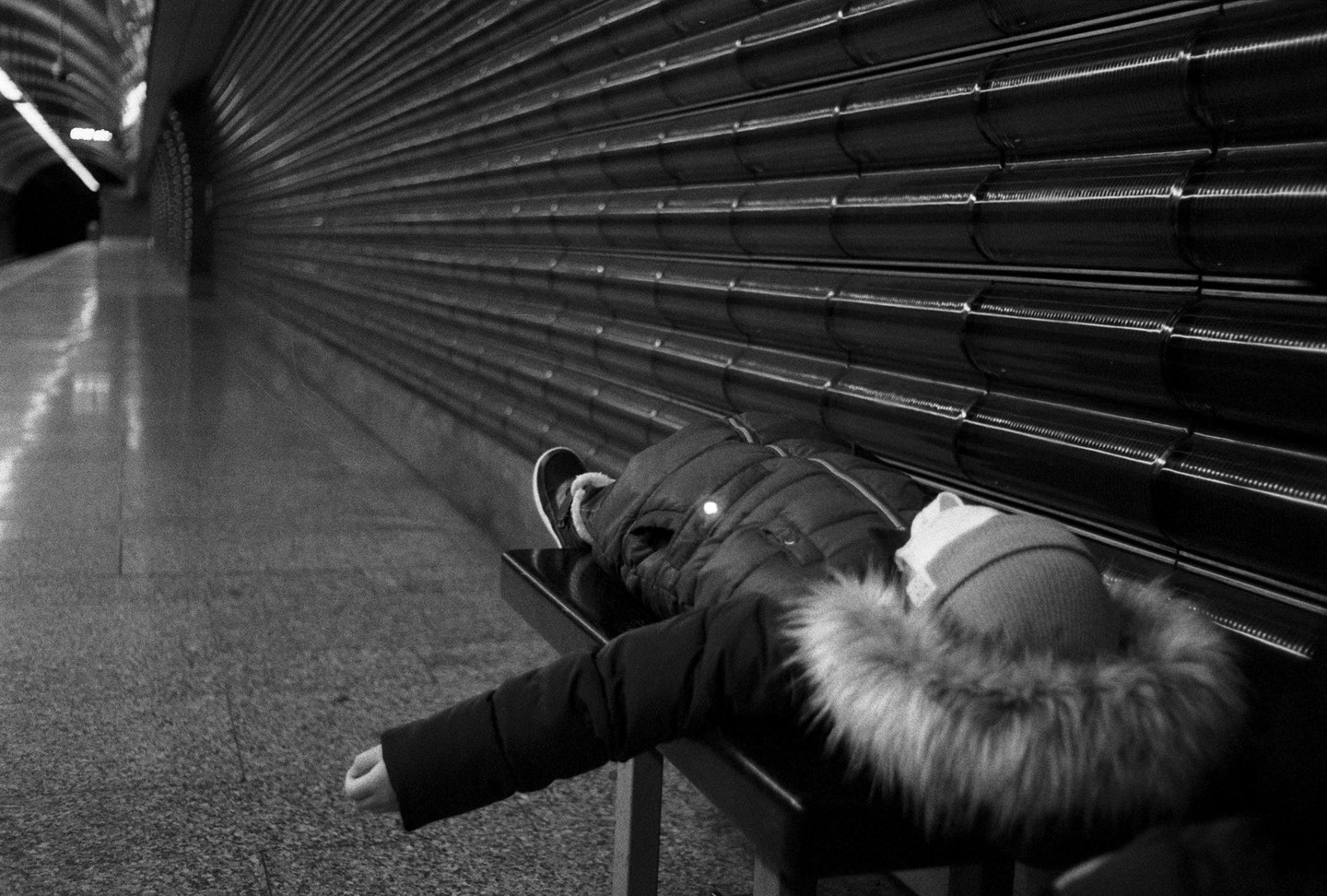 A person in a winter coat with a fur-lined hood lies on a bench in a subway station next to a textured wall.