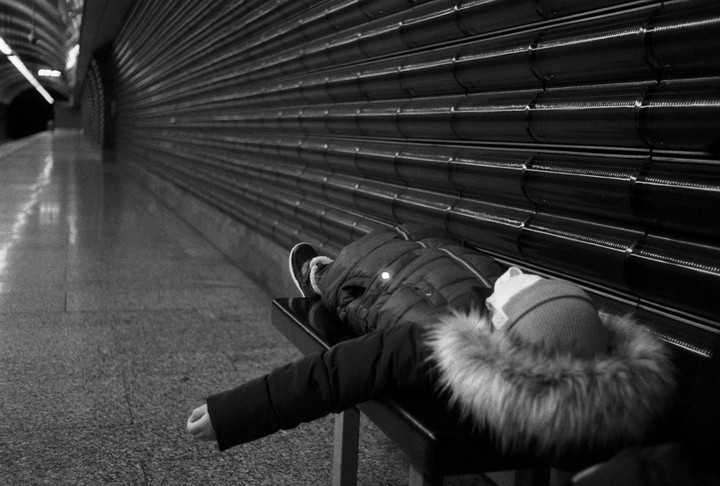 A person in a winter coat with a fur-lined hood lies on a bench in a subway station next to a textured wall.