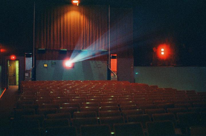 A projector beam shines onto a movie screen in an empty theater with rows of red seats.