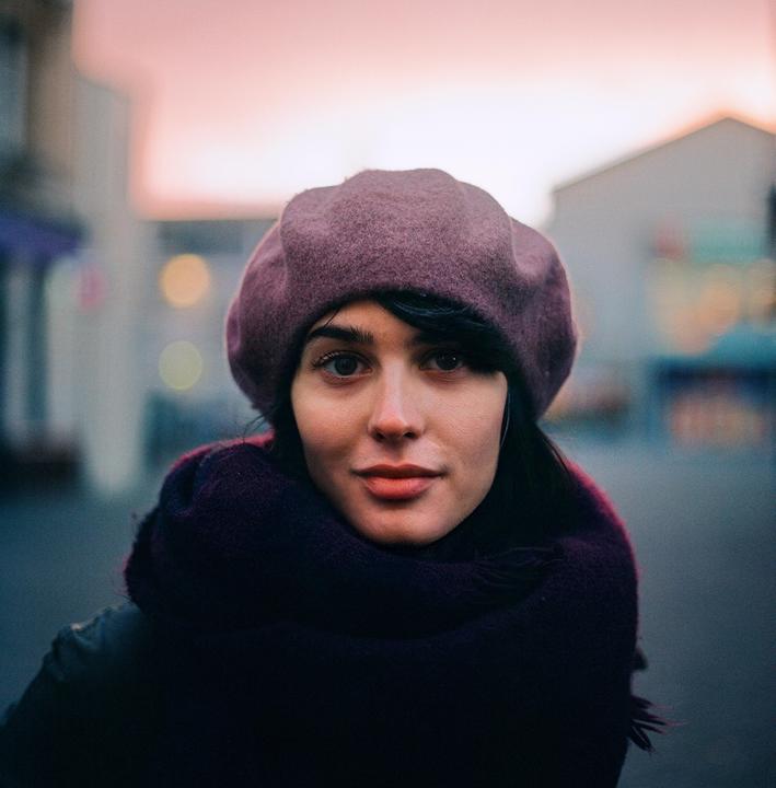 A woman wearing a beret and scarf looks directly at the viewer with a blurred street and pink sky in the background.