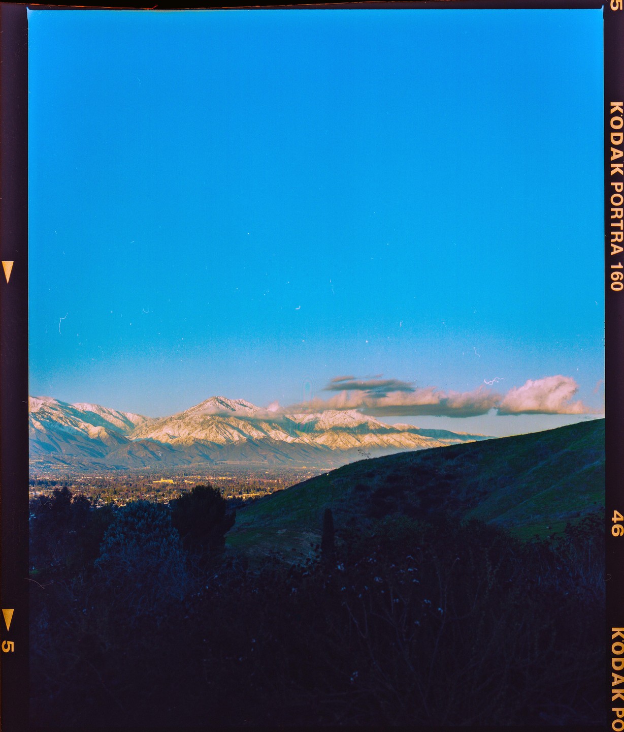 A wide landscape shows a valley with a city, surrounded by mountains with snow-capped peaks and a clear blue sky.