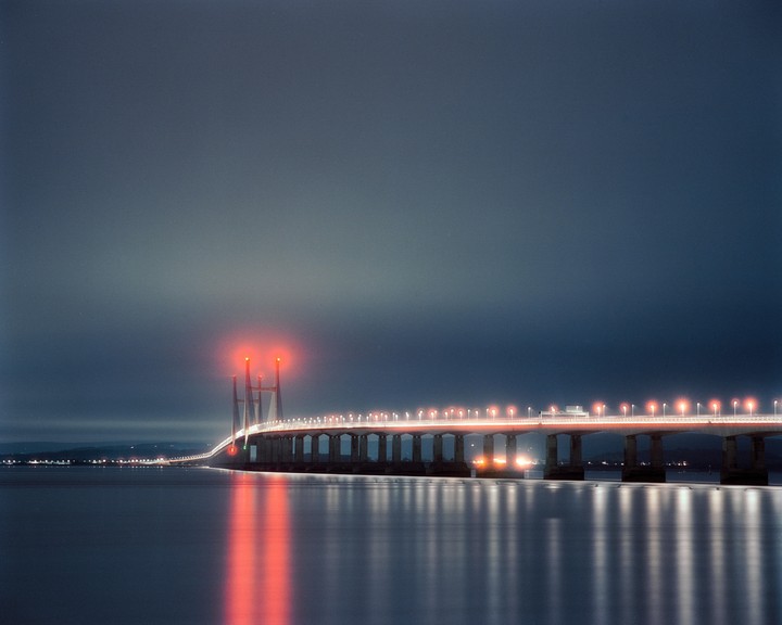 A long exposure shot of the Severn Bridge at night shows red and white lights reflecting on the water.