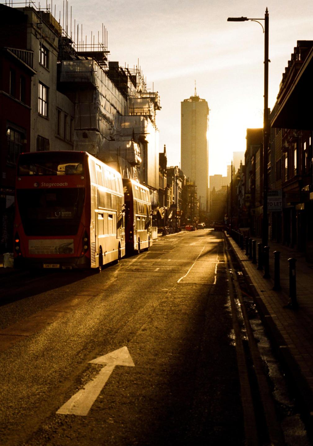 Two red double-decker buses are parked on a street with buildings under construction and a tall skyscraper in the background during golden hour.