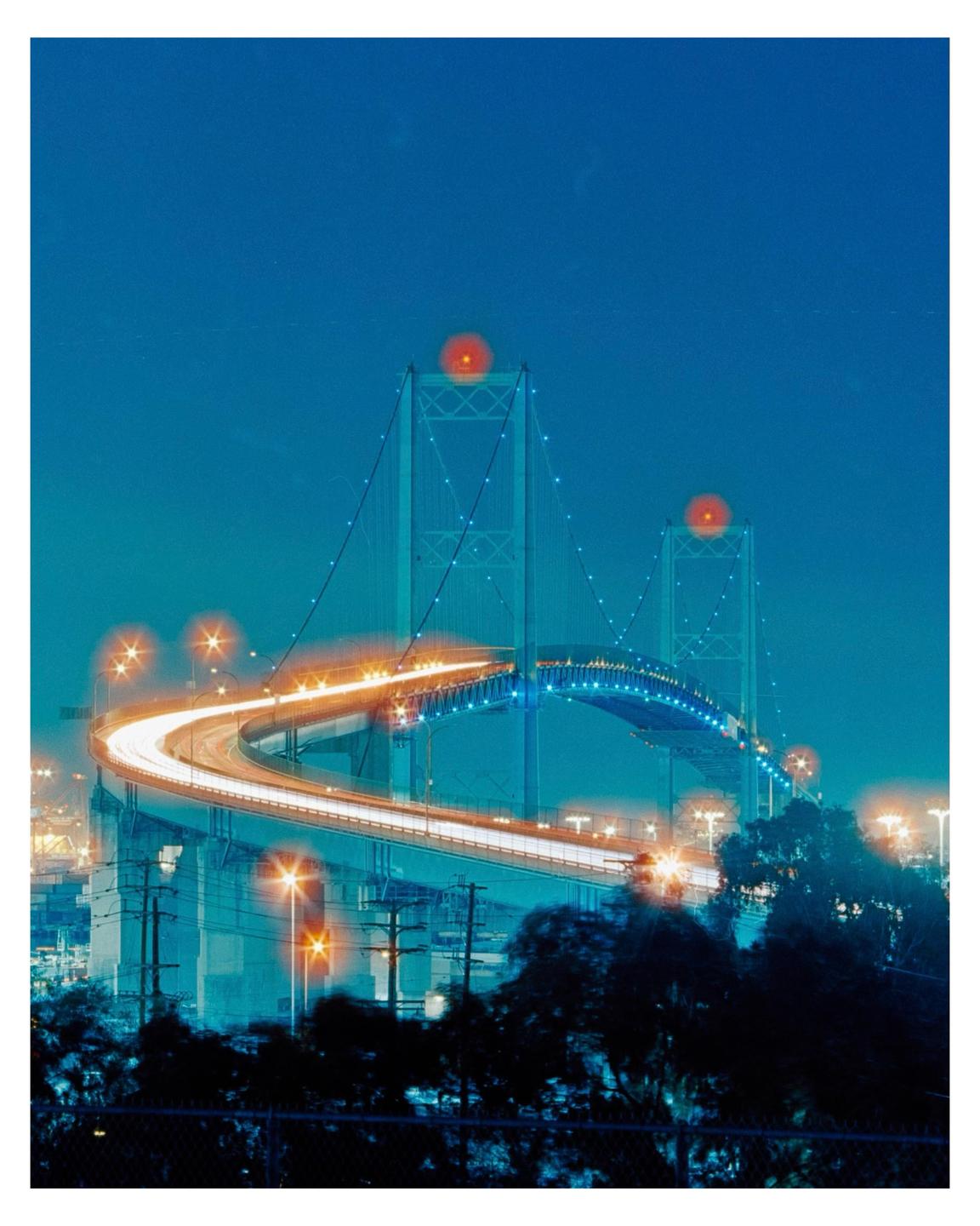 A long exposure shot captures the Vincent Thomas Bridge at night, with light trails from vehicles on the road.