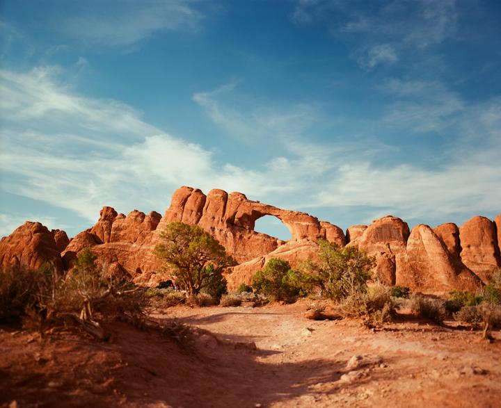 A natural arch is visible in the red rock formations of Arches National Park under a blue sky with wispy clouds.