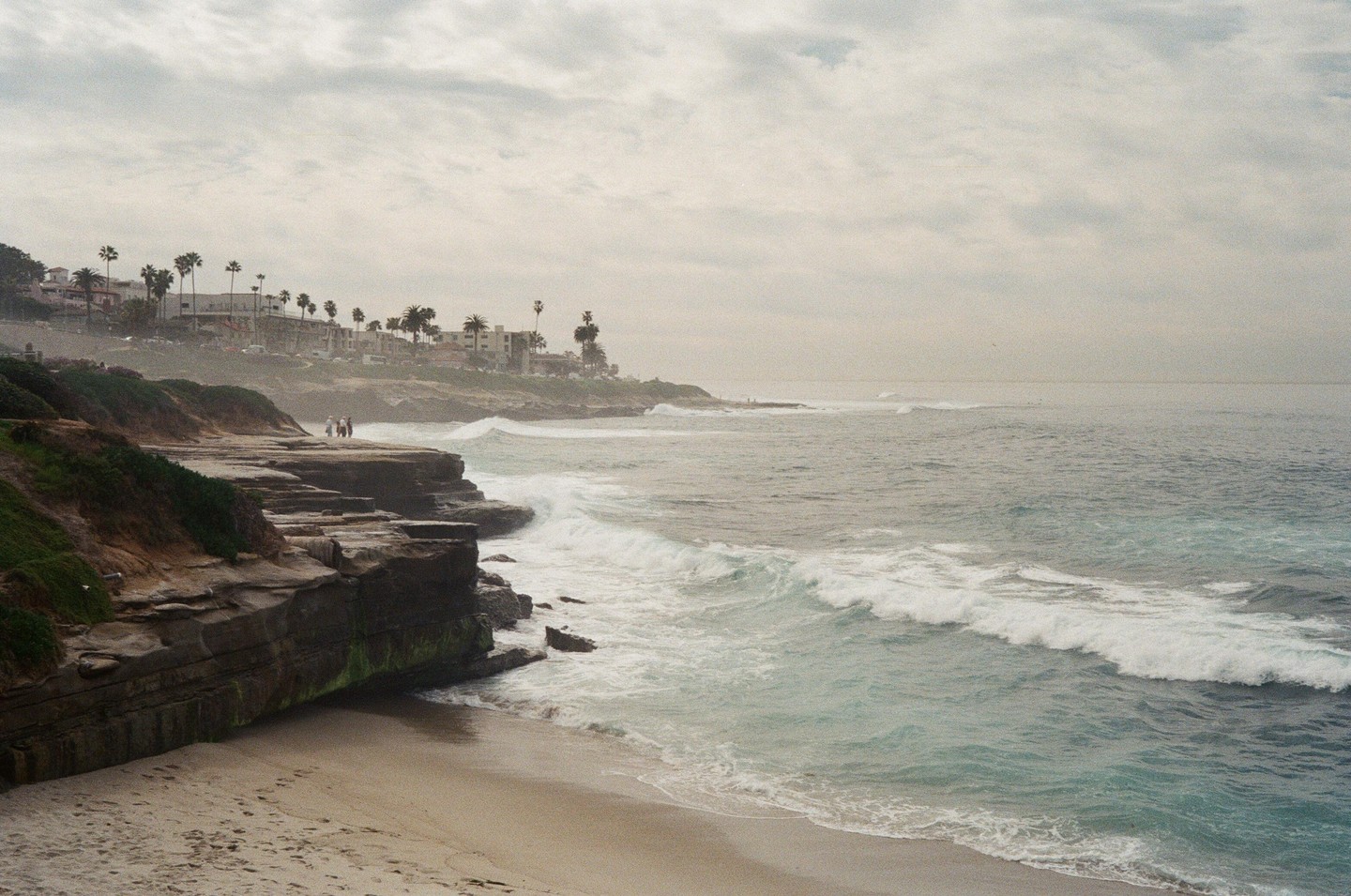 Waves crash against a sandy beach and rocky cliffs with buildings and palm trees in the background under a cloudy sky.