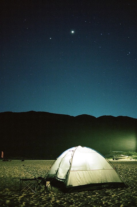 A white tent is illuminated on a sandy beach under a starry night sky with mountains in the background.