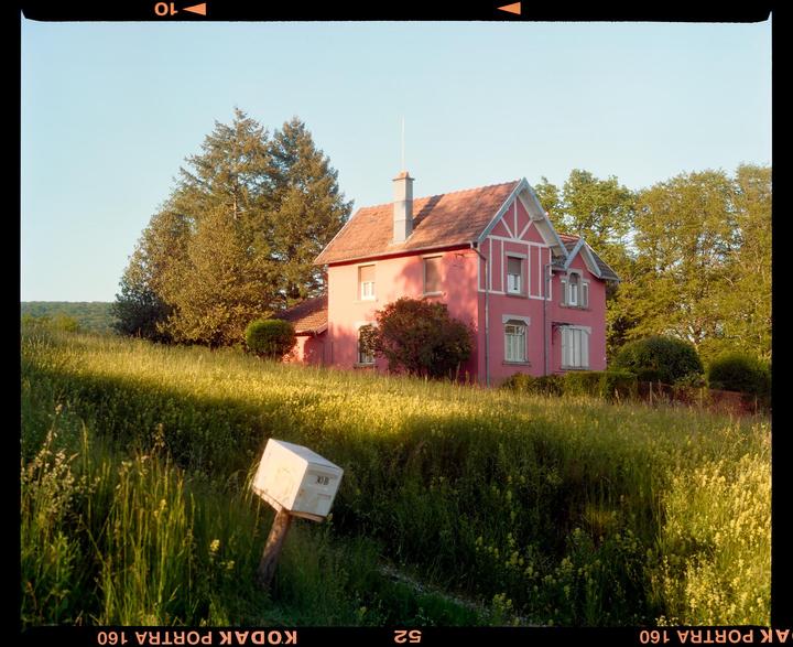 A pink house with a red roof stands on a hill surrounded by tall grass and trees.