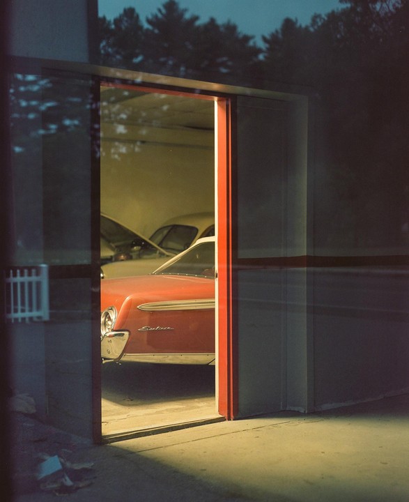 A red vintage car is parked inside a garage with its hood open, viewed through a glass door.