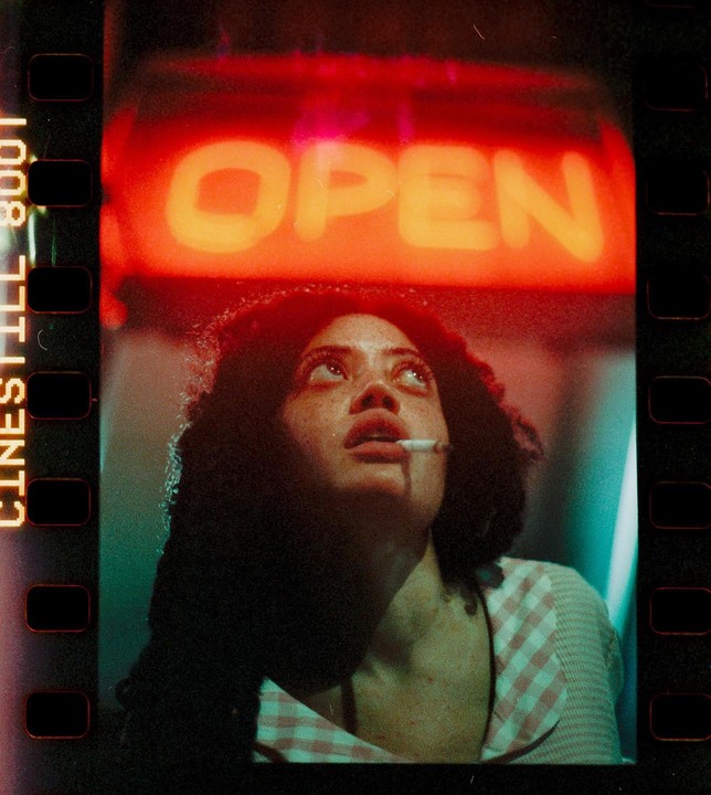 A woman with a cigarette in her mouth looks up at a neon "OPEN" sign.