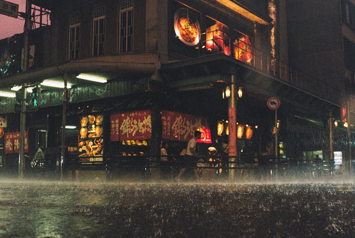 Rain falls on a street in front of a brightly lit building with red banners and lanterns at night.