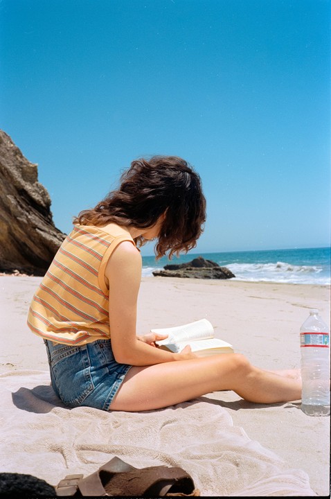 A woman sits on a towel on a sandy beach, reading a book with the ocean and cliffs in the background.