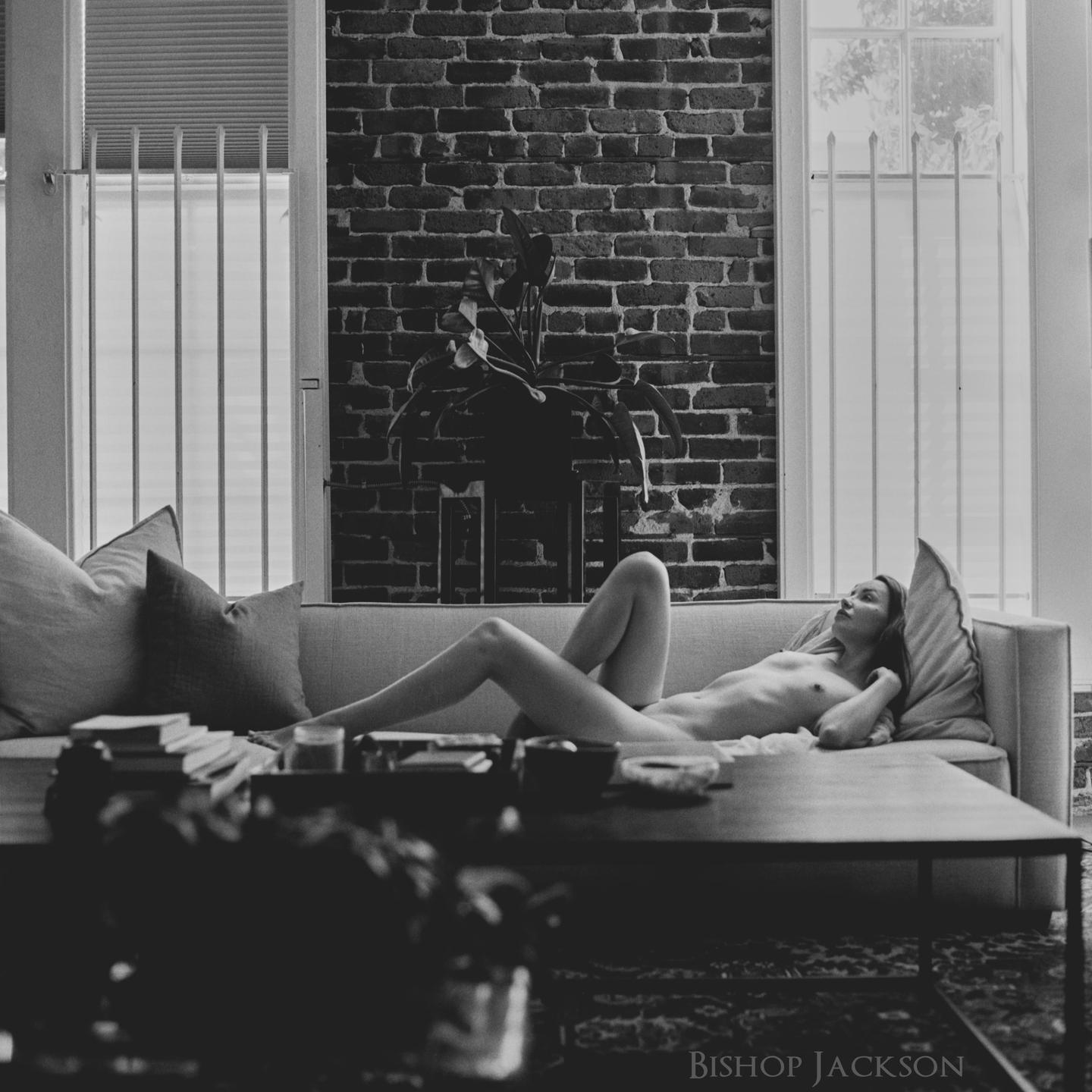 A nude woman reclines on a sofa in a room with a brick wall and windows.