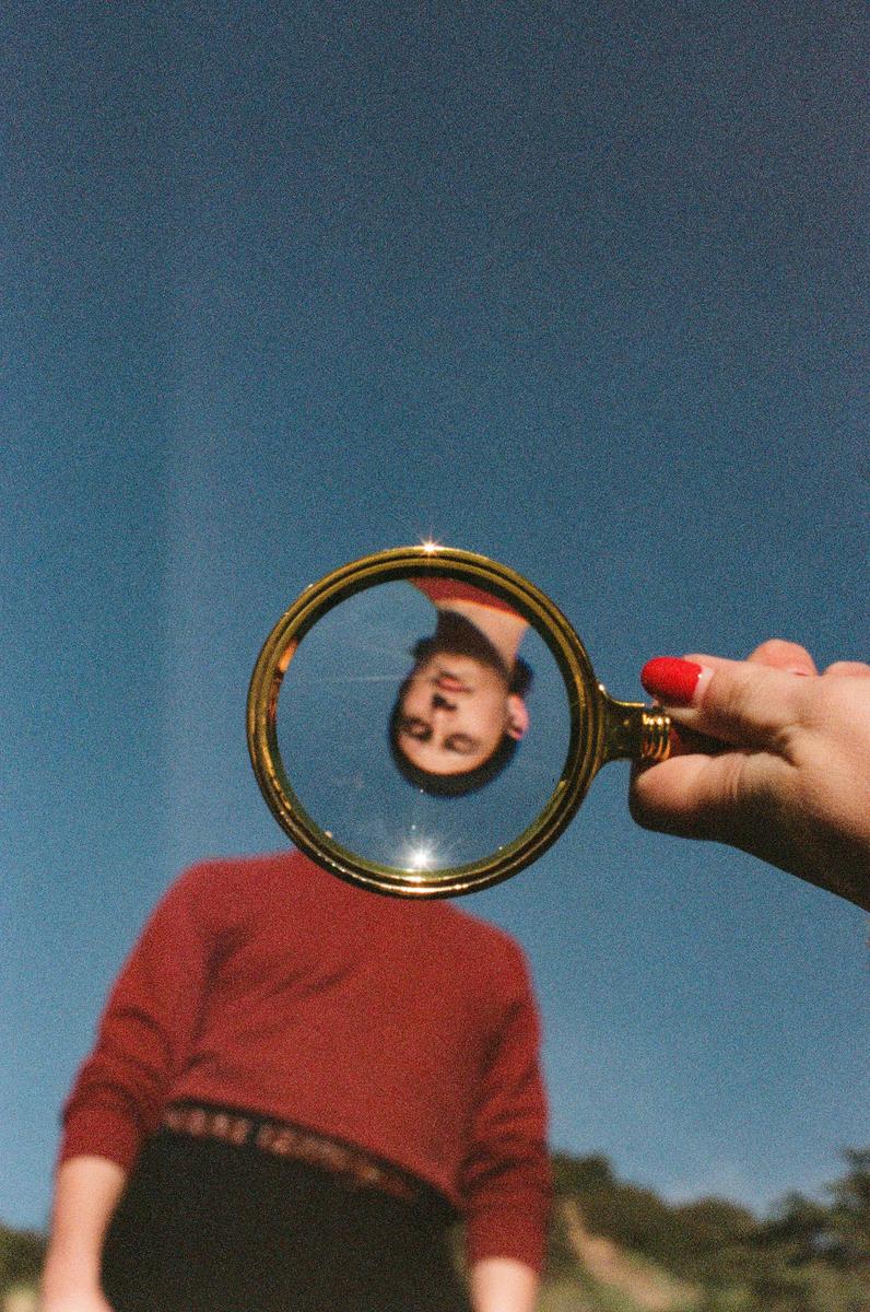 A person holds a magnifying glass that reflects an upside-down portrait of a man against a blue sky.