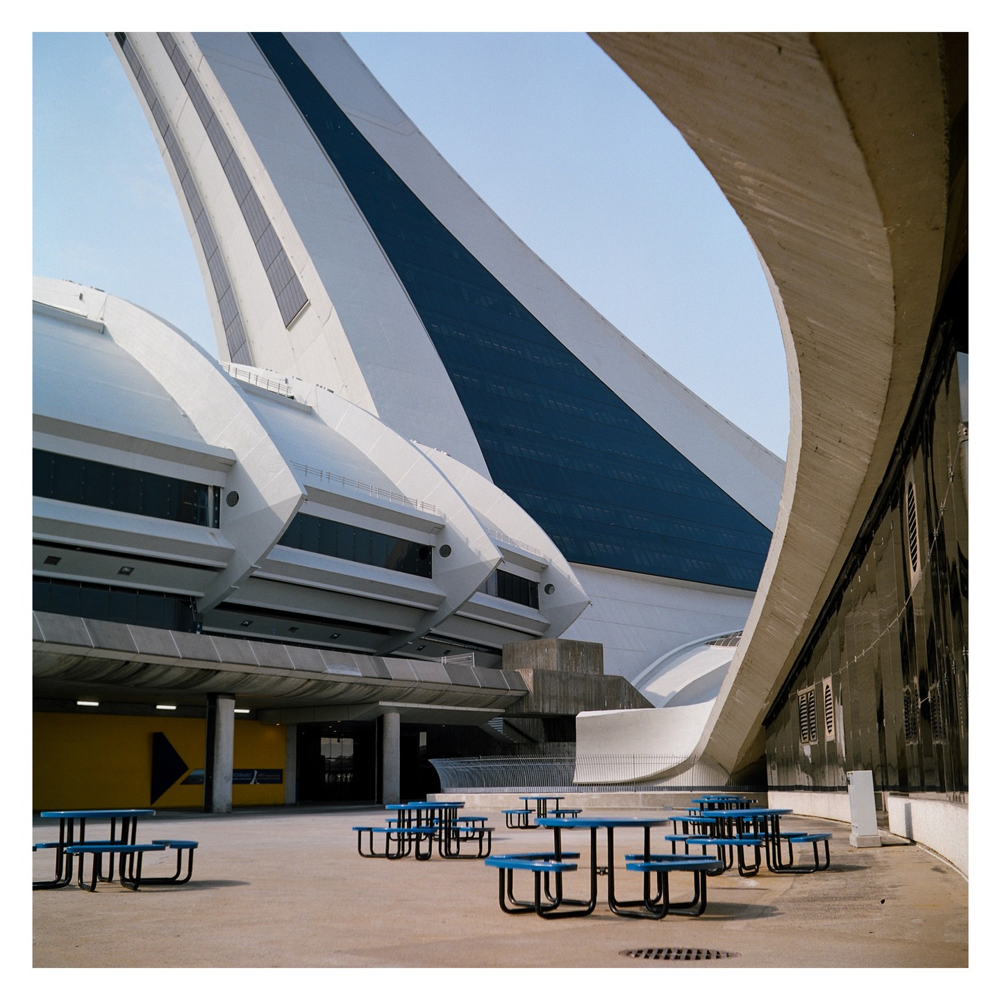 A large, modern stadium with a white and blue facade stands behind several blue picnic tables on a paved area.