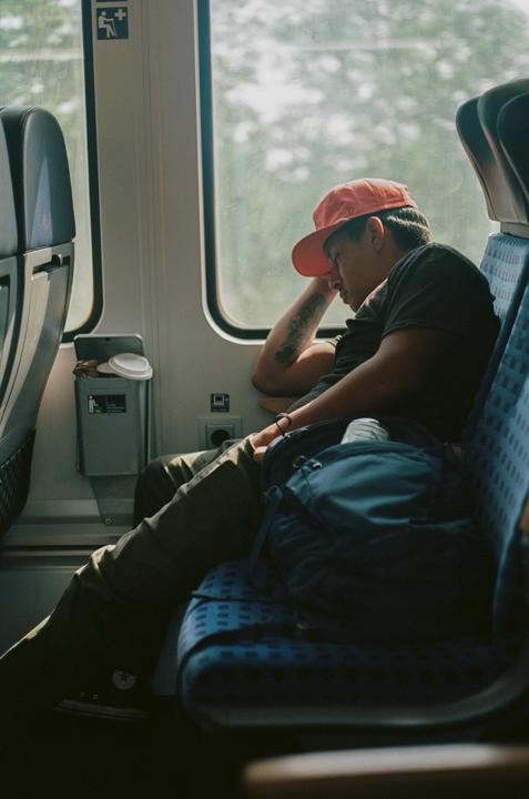 A man wearing a red cap is sleeping on a train next to a window.