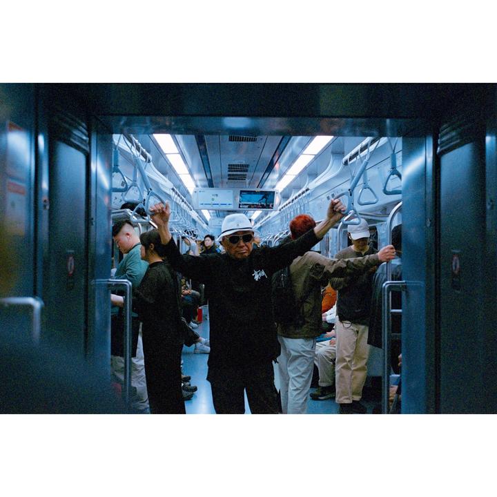 A man in sunglasses and a white hat stands with his arms raised in a subway car full of people.