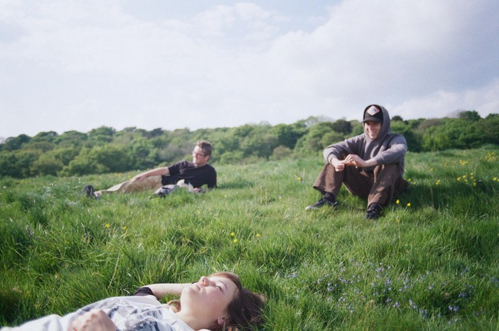 Three people are relaxing in a grassy field with trees in the background under a cloudy sky.