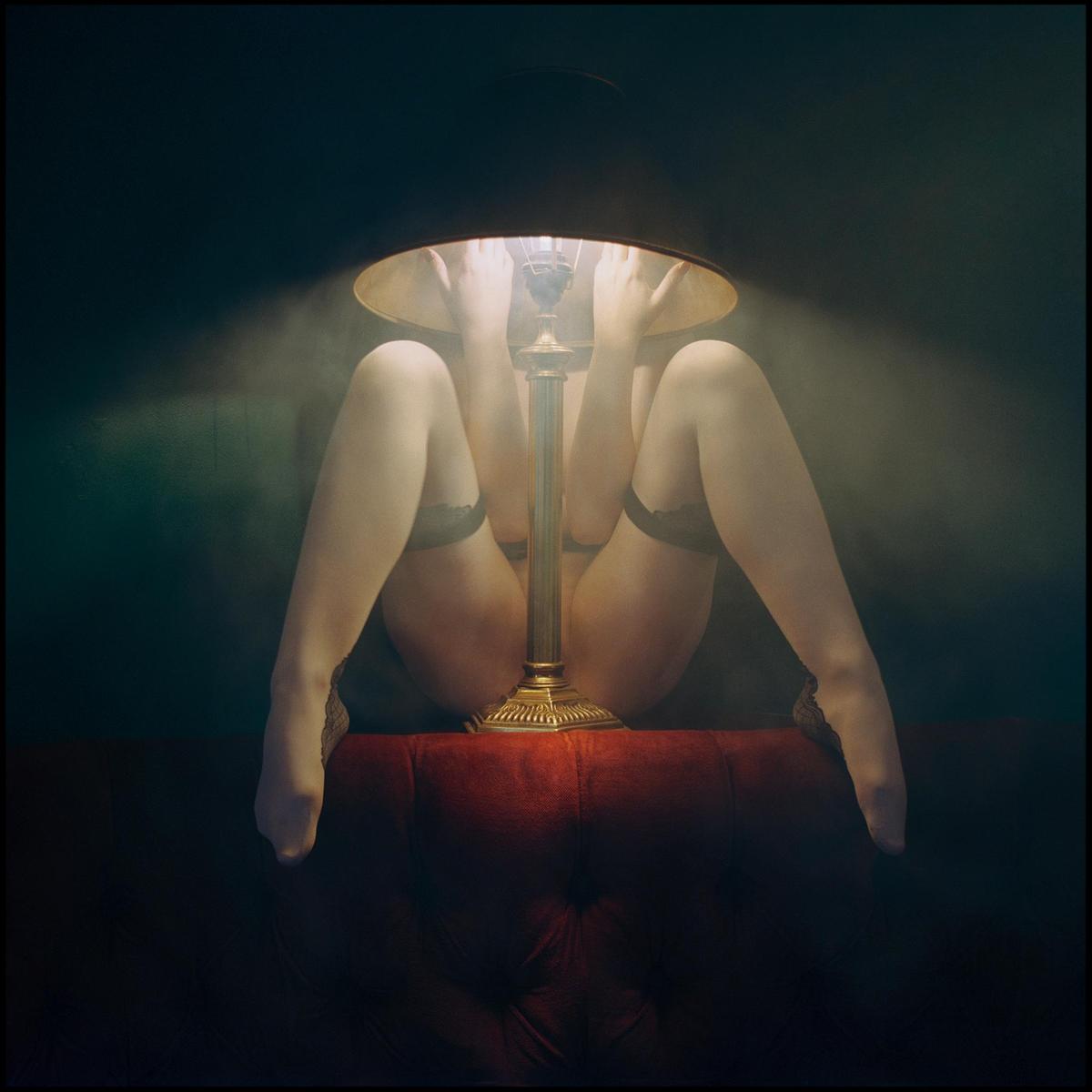 A woman's legs and hands are visible around a lit table lamp on a red tufted surface.