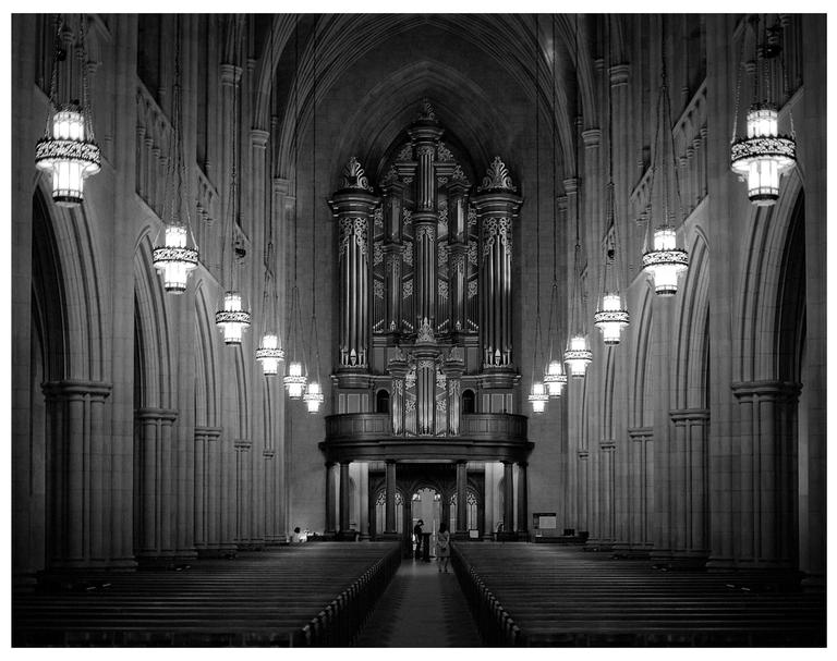A large pipe organ stands at the end of a long aisle in a church with rows of pews and hanging lights.
