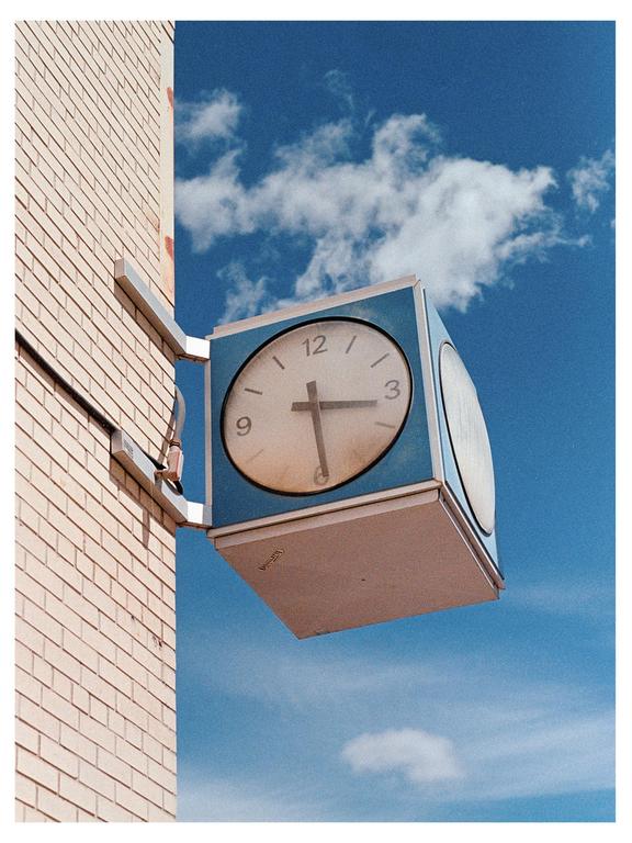 A square clock with a round face is mounted on a brick building against a blue sky with white clouds.