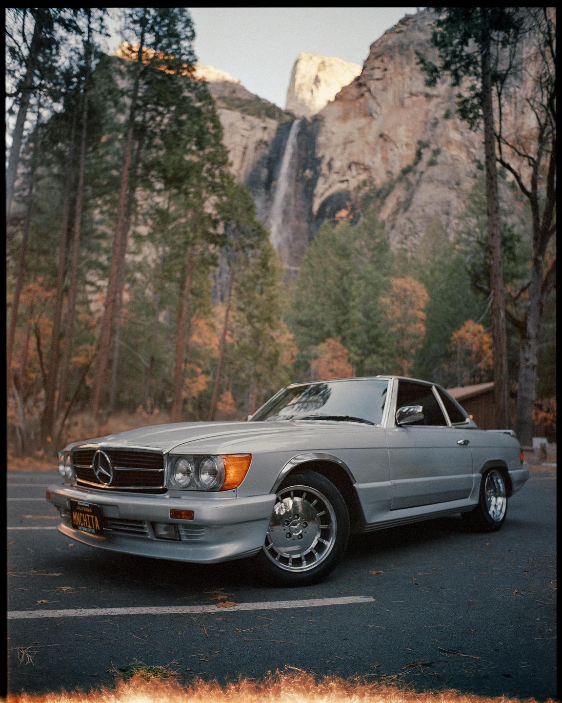 A silver Mercedes-Benz is parked on a road with a waterfall and mountains in the background.