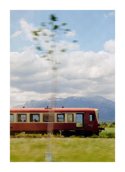 A red and white train travels through a green field with snow-capped mountains in the background under a cloudy sky.