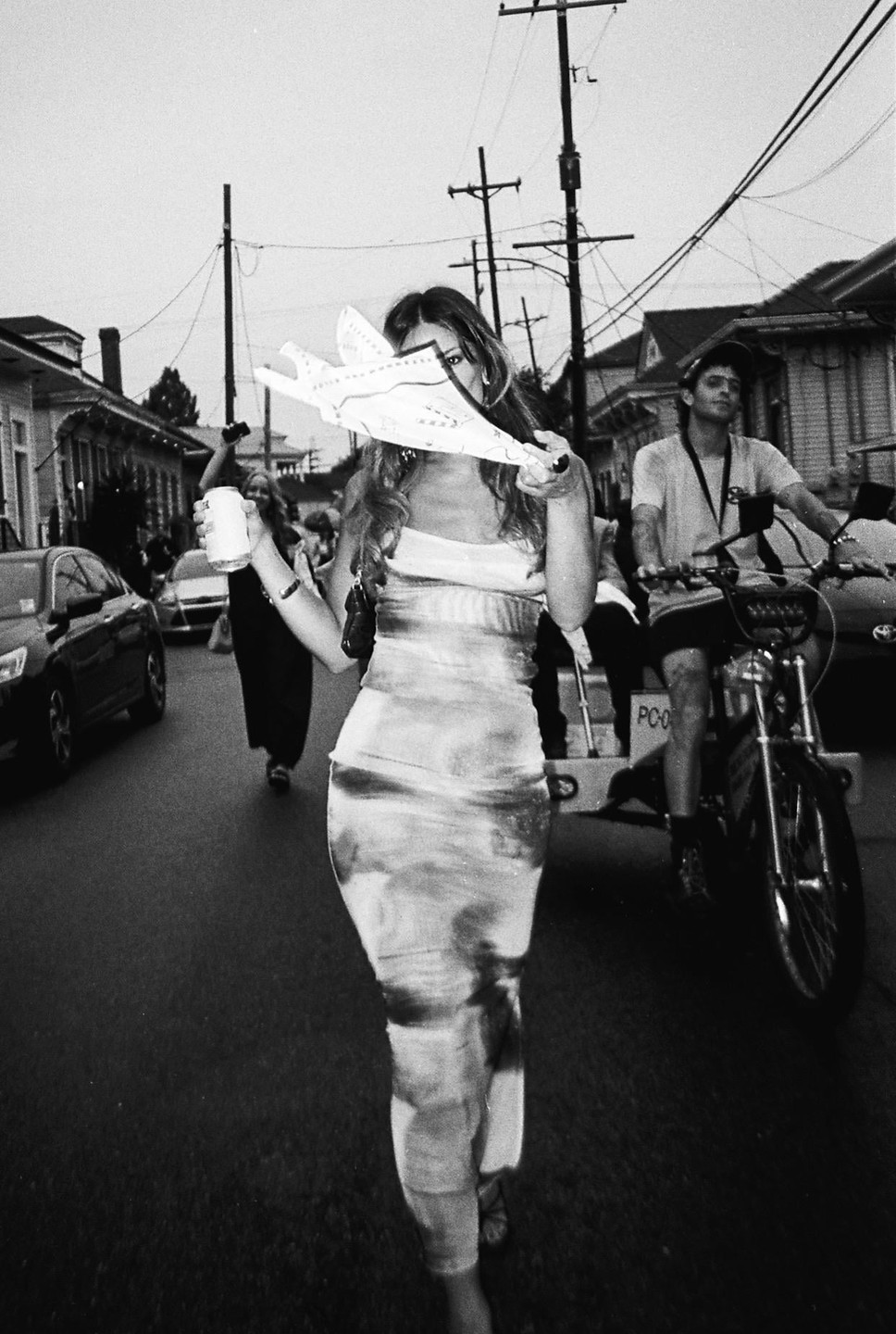 A woman in a dress holds a drink and a paper airplane in the street with other people and vehicles.