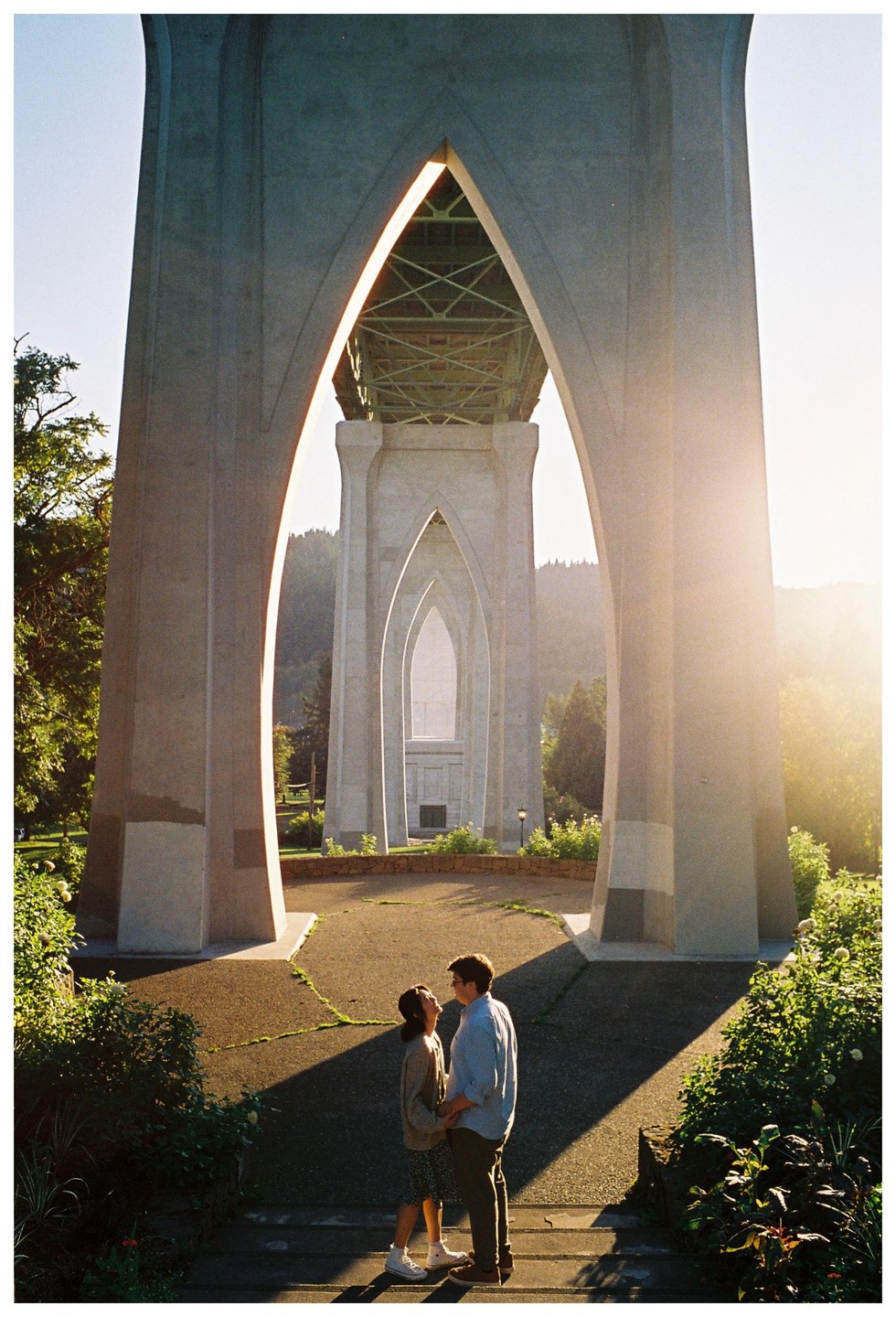 A couple stands holding hands under the arches of a large bridge at golden hour.