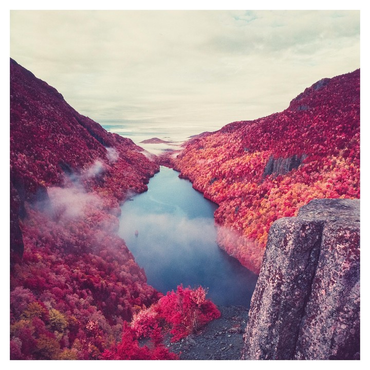 A body of water is nestled between two mountains covered in red and orange foliage, with fog in the valleys.