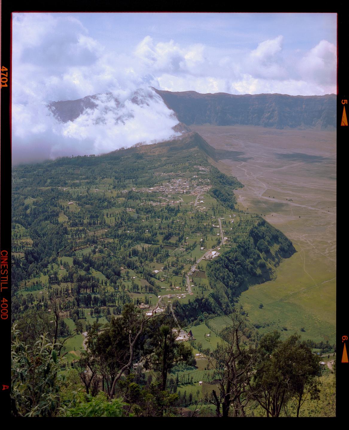 A village is nestled among green hills and trees, with a smoking volcano and a vast plain in the background under a cloudy sky.