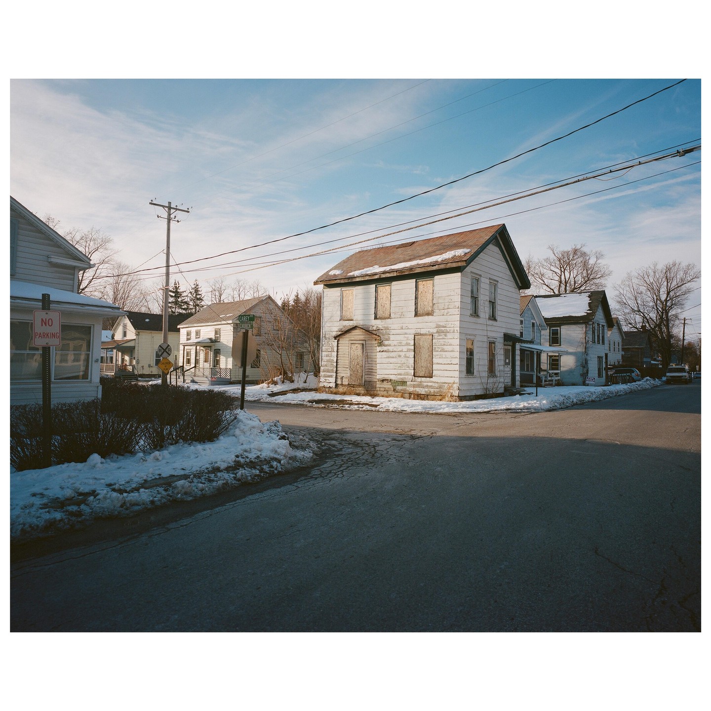 A white house with boarded-up windows stands at a snowy street corner under a blue sky.