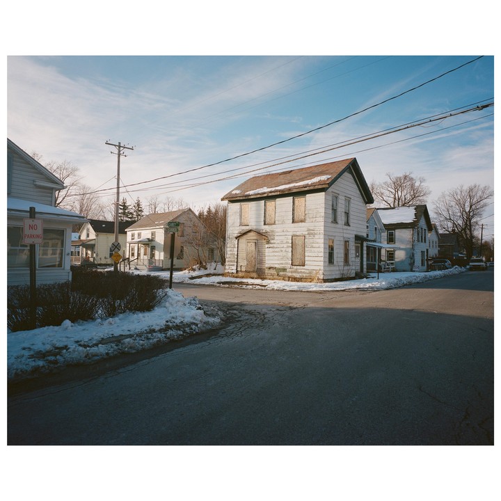 A white house with boarded-up windows stands at a snowy street corner under a blue sky.