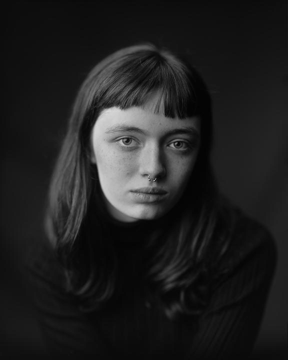 A woman with bangs and a septum piercing looks directly at the viewer.