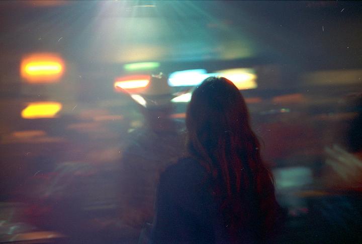 A woman with long hair stands in a dimly lit bar with colorful lights in the background.