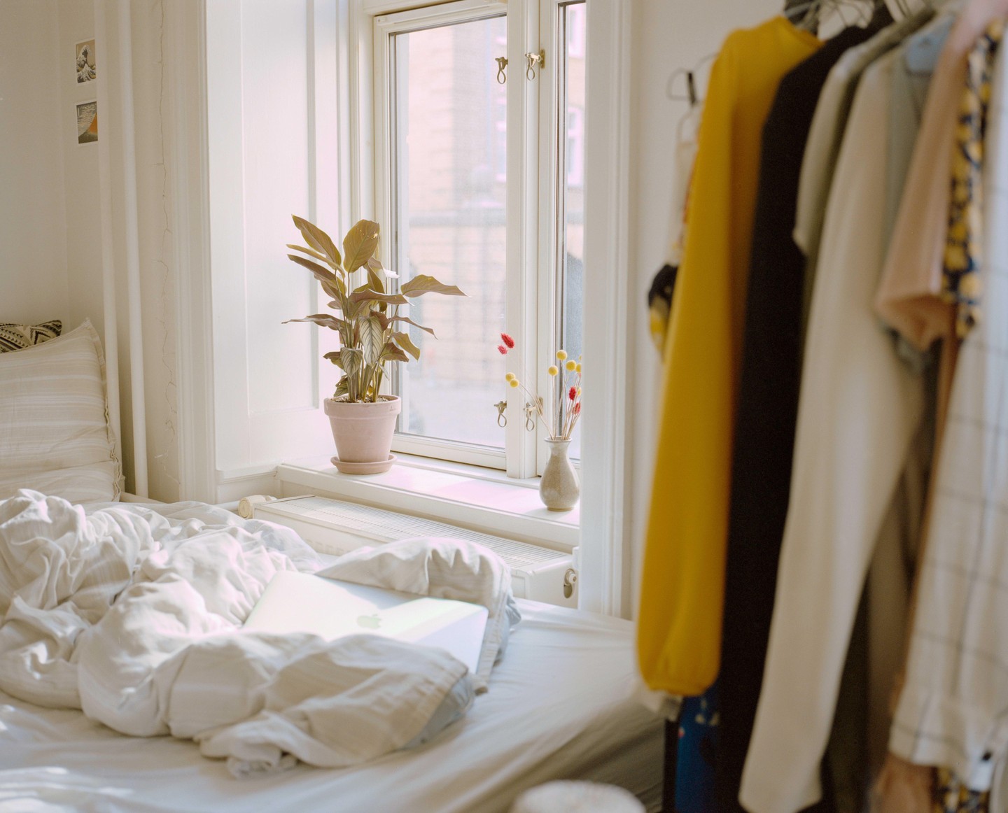 A sunlit bedroom features a bed with a laptop, a plant on the windowsill, and clothes hanging on a rack.