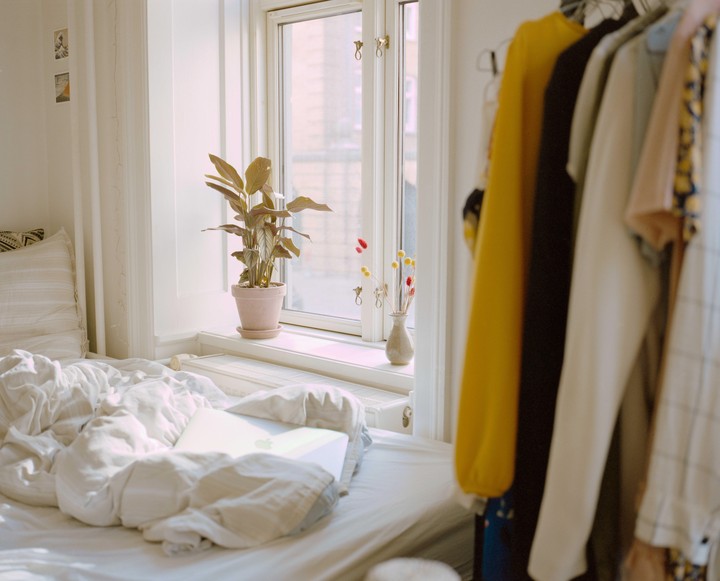 A sunlit bedroom features a bed with a laptop, a plant on the windowsill, and clothes hanging on a rack.